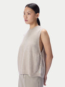 A woman shown in side profile wearing a sleeveless, cropped knit top in a light oatmeal beige color with a button detail along the side, paired with matching trousers, against a solid light gray background.
