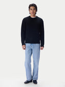 A man modeling a black cable-knit crewneck sweater, paired with light blue jeans and black loafers, against a plain white studio background.