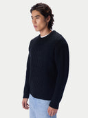 A man wearing a dark navy cable-knit crewneck sweater and light blue jeans, photographed from the side against a light gray studio background.