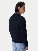 A man wearing a dark navy cable-knit sweater and light blue jeans, shown from a side-back angle against a plain light gray studio background.