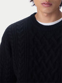 Close-up of a person wearing a black cable-knit sweater over a white undershirt, against a plain light background.