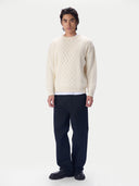 A man modeling a cream cable-knit crewneck sweater, paired with dark navy trousers and black shoes, against a plain white studio background.