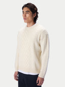 A man wearing a cream-colored cable-knit crewneck sweater, paired with dark trousers, photographed against a solid light gray background.