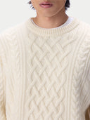 A close-up of a person wearing a cream-colored cable-knit sweater over a white undershirt, showing the detailed knit pattern against a light background.