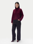 A model stands against a light gray studio background wearing a cropped, long-sleeve high-neck sweater in deep burgundy with a buttoned front, paired with black wide-leg trousers and burgundy pointed heels.