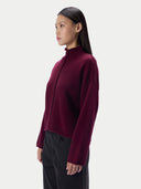 A woman shown in side profile wearing a cropped, long-sleeve knit top in deep burgundy with a braided front detail, paired with dark pants, against a light gray studio background.