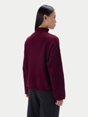 A woman shown from the back wearing a dark burgundy mock-neck knit sweater and black pants, photographed against a clean light gray studio background.