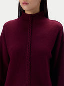 A close-up of a model wearing a deep burgundy knit sweater with a high mock neck and a central braided detail running down the front.