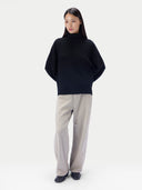 A model stands against a light gray studio background wearing a black oversized mock-neck knit sweater, light beige wide-leg trousers, and black shoes.