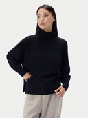 A woman modeling a black turtleneck sweater with a relaxed, oversized fit and light gray trousers, set against a plain light gray studio background.