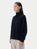 A woman wearing a black ribbed mock neck sweater and light beige trousers, photographed from the side against a light gray studio background.