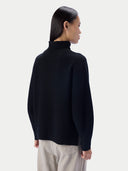 A woman shown from the back wearing a black turtleneck sweater and light beige trousers, photographed against a clean light gray studio background.
