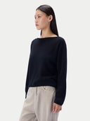 A woman wearing a black boat-neck knit sweater and light beige trousers, photographed against a clean light gray studio background.