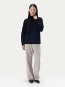 A woman wearing a black loose-fit turtleneck sweater, light gray wide-leg trousers and black flat shoes, standing against a light gray studio background.