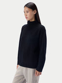 A woman shown in three-quarter profile wearing a black turtleneck knit sweater with ribbed cuffs and hem and light gray trousers, against a light gray studio background.
