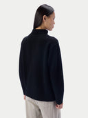 A woman shown from the back wearing a black turtleneck knit sweater with a ribbed collar and light gray trousers, against a light gray studio background.