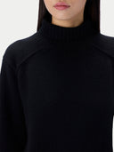 A close-up of a woman wearing a black knit sweater, showing the ribbed turtleneck collar and raglan sleeve seam.