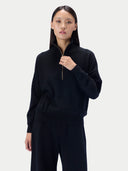 A model wearing a black long-sleeve quarter-zip pullover sweater and matching black pants, standing against a light gray studio background.