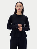 A woman modeling a black long-sleeve knit top with a peplum hem and button cuffs, paired with black pants, against a light gray studio background.