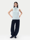 A model stands against a light gray studio background wearing a sleeveless light blue mock-neck knit top and dark wide-leg trousers.