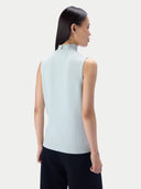 A woman shown from the back wearing a sleeveless mock-neck knit top in a very light icy blue color, paired with dark pants, against a plain light gray studio background.