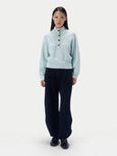 A model stands against a light gray studio background wearing a pale icy blue mock-neck knit sweater with three front buttons, paired with dark wide-leg trousers and black shoes.