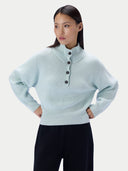 A woman modeling a light mint green ribbed knit mock-neck sweater with a buttoned henley neckline, paired with dark pants, against a plain light gray studio background.