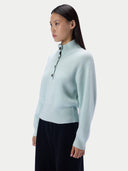 A woman wearing a pale mint ribbed mock-neck sweater with a short button placket and dark pants, photographed against a clean light gray studio background.