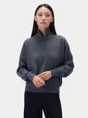 A woman modeling a dark gray heathered quarter-zip knit sweater with a stand collar, paired with black ribbed pants, against a light gray studio background.