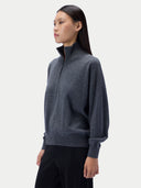A woman modeling a dark gray quarter-zip knit sweater with a mock neck, paired with black pants, against a light gray studio background.