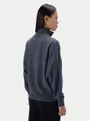 A woman shown from the back wearing a dark charcoal gray mock neck sweater and black trousers, photographed against a clean light gray studio background.