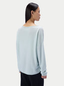 A woman shown from the back wearing a pale icy blue long-sleeve knit top and black pants against a light gray studio background.