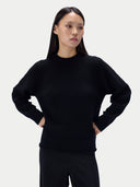 A woman modeling a black long-sleeve knit sweater with a crew neck, paired with black ribbed pants, against a light gray studio background.