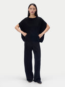 A model stands against a light gray studio background wearing a black short-sleeve knit top with draped sleeves and matching black wide-leg trousers.