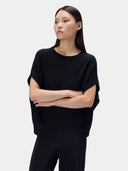 A model wearing a black short-sleeve knit top with draped, wide sleeves and black ribbed trousers, posed against a light gray studio background.
