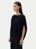 A woman modeling a black knit top with wide draped sleeves and side cutouts, paired with black pleated trousers, against a light gray studio background.
