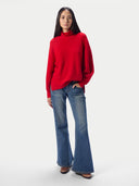 A woman modeling a bright red mock-neck knit sweater and blue flare jeans against a plain light gray studio background.