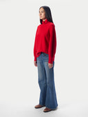 A woman modeling a bright red long-sleeve turtleneck sweater paired with blue wide-leg jeans and brown shoes, standing against a plain light gray studio background.