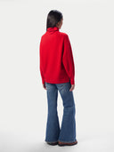 A model shown from the back wearing a bright red turtleneck sweater and blue flared jeans against a plain light gray studio background.