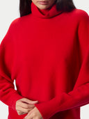 A close-up of a model wearing a bright red, oversized turtleneck sweater against a plain light background.
