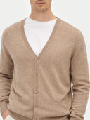 A man wearing a light brown knit V-neck cashmere cardigan over a white t-shirt, photographed against a solid light gray background. #color_taupe