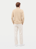 A man viewed from behind wearing a beige mock-neck knit sweater, off-white trousers, and white sneakers against a light gray studio background.