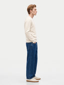 A man in profile wearing a cream knit sweater, blue wide-leg jeans, and cream leather shoes, standing against a plain light gray studio background.