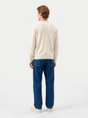 A man viewed from behind wearing a cream-colored knit sweater and blue jeans against a plain light gray studio background.