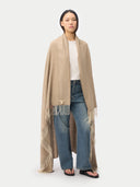 A model stands against a light gray studio background wearing a long beige fringed wrap or shawl over a white top and blue wide-leg jeans. #color_off-white