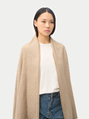 A model stands against a light gray studio background wearing a long beige fringed wrap or shawl over a white top and blue wide-leg jeans. #color_off-white