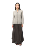 A woman stands against a plain white background wearing a light gray zip-front cardigan with a stand collar and a long dark brown skirt.