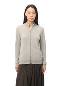 A woman modeling a light gray zip-up cardigan sweater with a stand-up collar, paired with dark pleated trousers, against a plain white background.