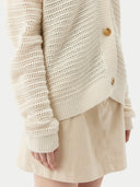 A woman wearing a beige cotton cashmere cardigan with a crew neckline and button closures over a black top and a beige skirt. #color_pure-white