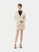 A woman wearing a beige cotton cashmere cardigan with a crew neckline and button closures over a black top and a beige skirt. #color_pure-white
